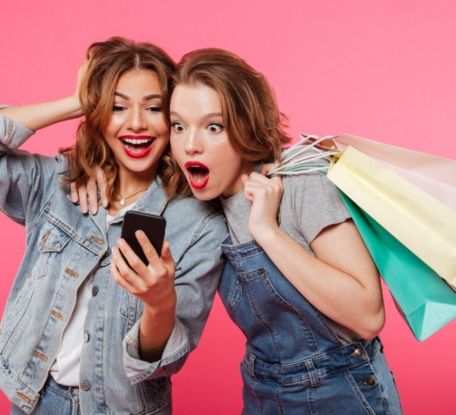 shocked-two-women-friends-holding-shopping-bags-using-mobile-phone